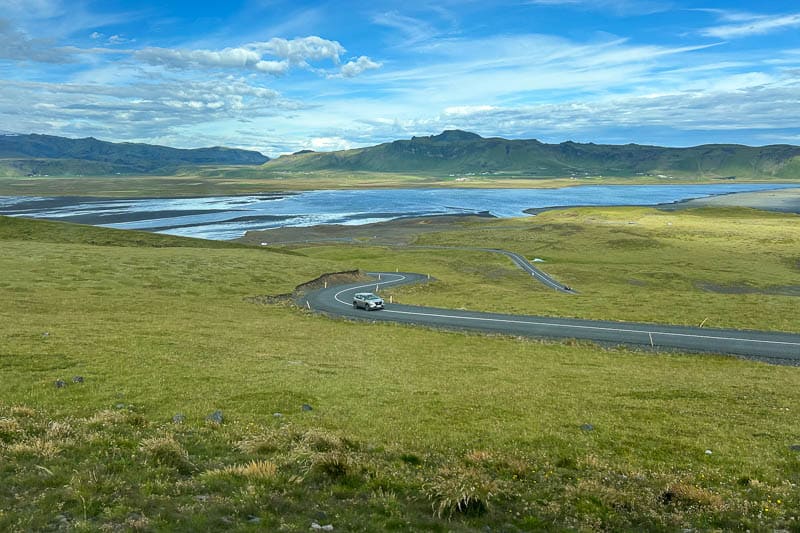 A car drives along a winding road through a green landscape with a river, mountains, and a blue sky with clouds—capturing the beauty of 10 days in Iceland.