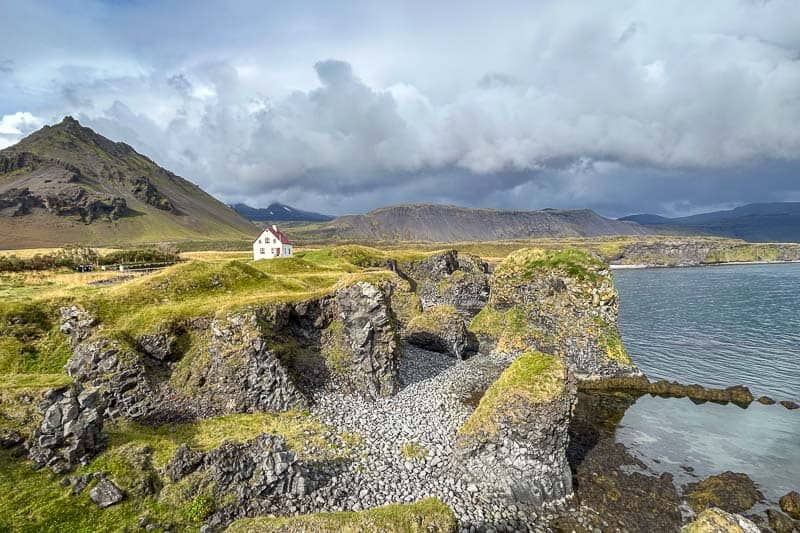 A white house with a red roof sits on a grassy hill above rocky cliffs and a pebble beach, framed by misty mountains and cloudy skies—an iconic scene for anyone going to Iceland near its dramatic coastlines.