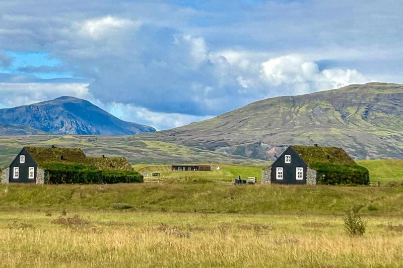 10 Days in Iceland: Our Real Itinerary 54 Two black houses with grass roofs sit in a grassy field, surrounded by rolling green hills and a mountain under a partly cloudy sky—a serene scene you might encounter during 10 days in Iceland.