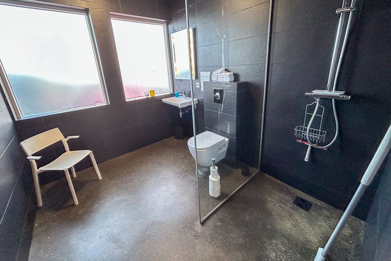 Kef Guesthouse Review - A Handy Overnight Stay Near Keflavík Airport 5 A modern, accessible Kef guesthouse bathroom with black walls, a wall-mounted toilet, basin, shower area with handheld showerhead, white chair, frosted windows, and a clear glass shower screen.