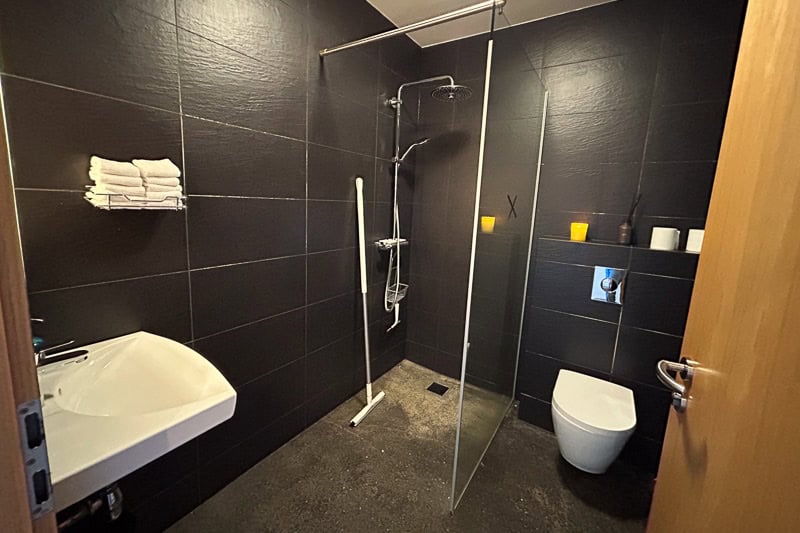 Kef Guesthouse Review - A Handy Overnight Stay Near Keflavík Airport 6 A modern bathroom at Kef guesthouse features dark tiles, a wall-mounted basin, a walk-in shower with a glass divider, and a wall-mounted toilet. Candles and toiletries rest on a shelf above the toilet, with towels stacked on a wall rack.
