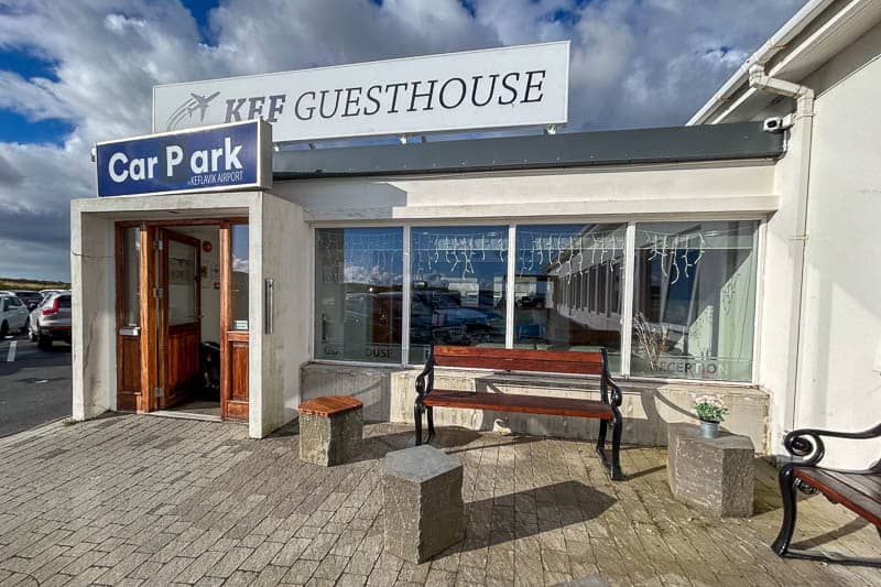 Exterior view of Kef Guesthouse with a sign above the entrance, a Car Park sign, wooden bench, and stone stools outside the front windows. The sky is partly cloudy, adding charm to Kef Guesthouse’s welcoming exterior.