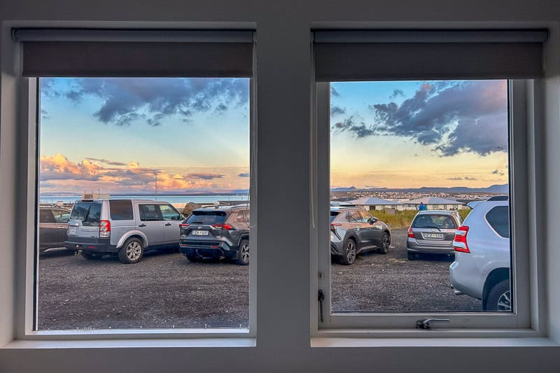 Kef Guesthouse Review - A Handy Overnight Stay Near Keflavík Airport 9 View through two windows at Kef Guesthouse reveals a car park with several cars on gravel, grassy area beyond, and a cloudy sky tinged with pinkish sunset hues.