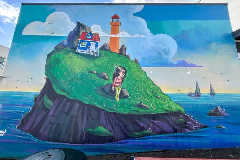 10 Days in Iceland: Our Real Itinerary 53 A mural inspired by 10 days in Iceland depicts a girl sitting on a grassy island with a house and lighthouse beside her, surrounded by ocean and distant rocks under a partly cloudy sky.