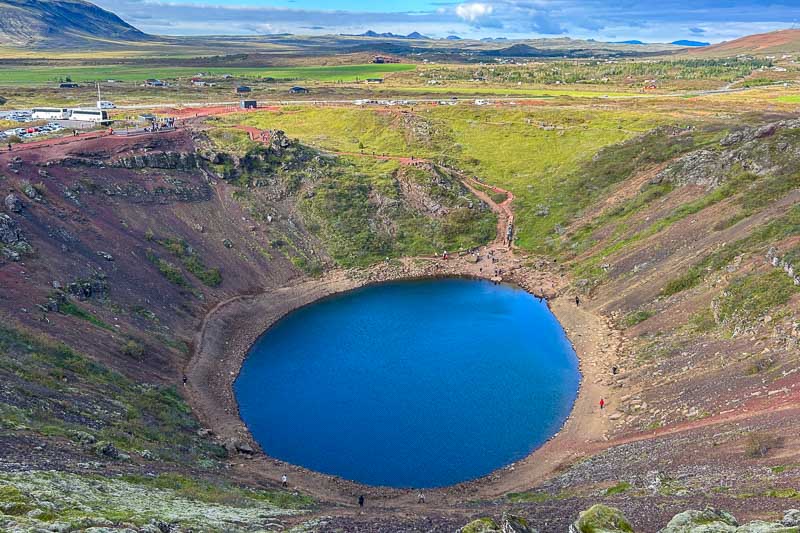A large, circular volcanic crater with deep blue water sits in a rocky landscape, surrounded by green fields and distant mountains—just one of the unforgettable sights you’ll see during 10 days in Iceland under a partly cloudy sky.