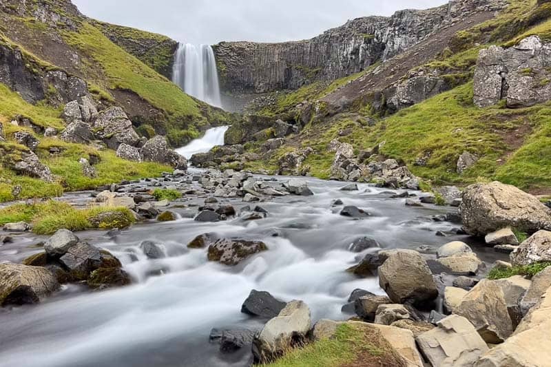 10 Days in Iceland: Our Real Itinerary 55 A waterfall cascades down a cliff into a rocky stream, surrounded by green hills and large boulders under a cloudy sky—just one of the many breathtaking sights you’ll encounter during 10 days in Iceland.