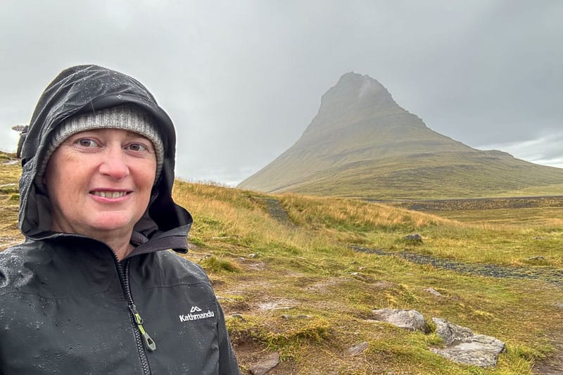10 Days in Iceland: Our Real Itinerary 50 A person wearing a rain jacket and beanie stands outside on a grassy landscape with a mountain in the background under cloudy skies, capturing the adventurous spirit of 10 days in Iceland.