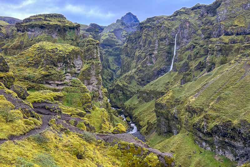 10 Days in Iceland: Our Real Itinerary 24 A deep green canyon with steep rock walls, a narrow river at the bottom, a tall waterfall on the right, and a cloudy sky overhead—an unforgettable sight during 10 days in Iceland.