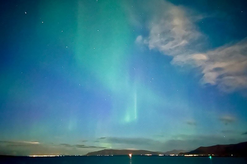 10 Days in Iceland: Our Real Itinerary 51 Northern lights with green and blue hues dance above a coastal landscape in Iceland, where mountains, clouds, and city lights line the horizon—a magical sight you might witness during 10 days in Iceland.