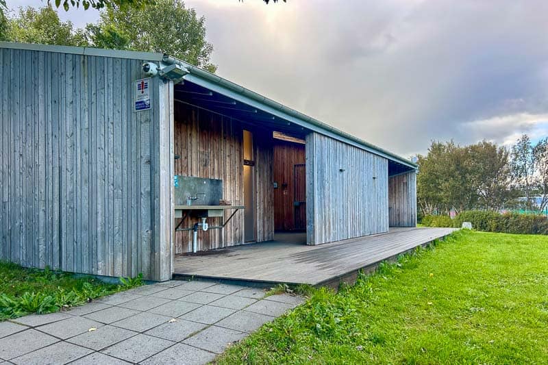 Iceland Campgrounds 17 A modern wooden building with an open doorway, metal sink, and covered porch sits next to a grassy lawn and trees under a cloudy sky.