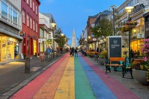 10 Days in Iceland: Our Real Itinerary 48 A city street, reminiscent of adventures like 10 days in Iceland, features rainbow-colored pavement lined with shops and cafes, leading to a tall church in the distance under a blue evening sky.