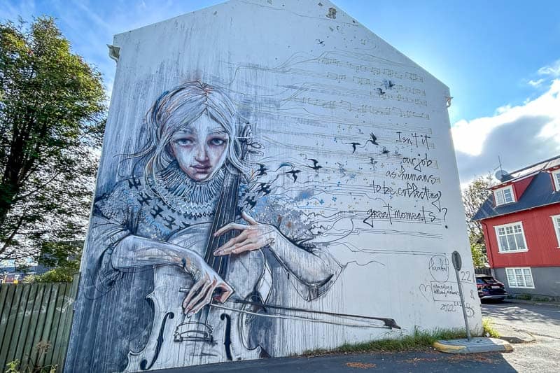 10 Days in Iceland: Our Real Itinerary 6 Large mural on a white building depicting a somber person playing a cello, with musical notes, birds, and handwritten text—evoking the reflective mood of 10 days in Iceland.
