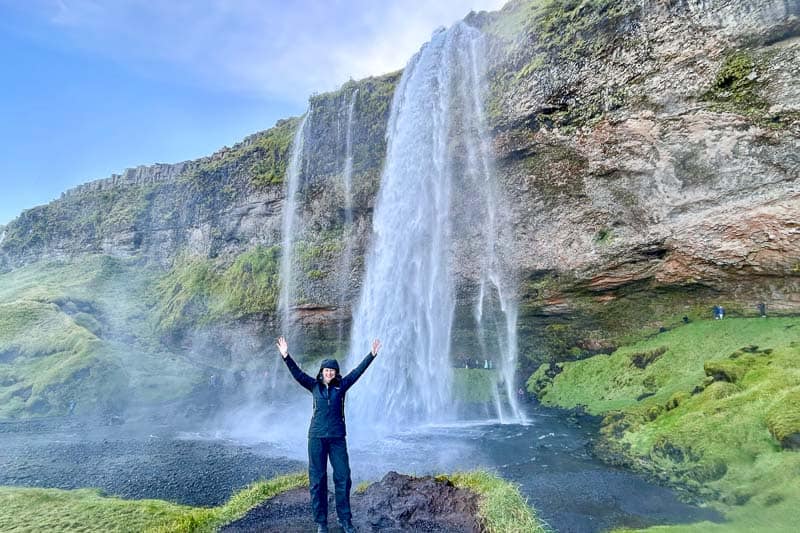 A person in dark clothing stands with arms raised in front of a tall waterfall cascading down a rocky, green cliff under a blue sky, capturing the adventure of 10 days in Iceland.