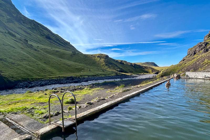 10 Days in Iceland: Our Real Itinerary 15 A rectangular outdoor swimming pool sits next to a grassy valley surrounded by green hills under a clear blue sky—an inviting spot to relax during your 10 days in Iceland, as a few people enjoy bathing in the pool.