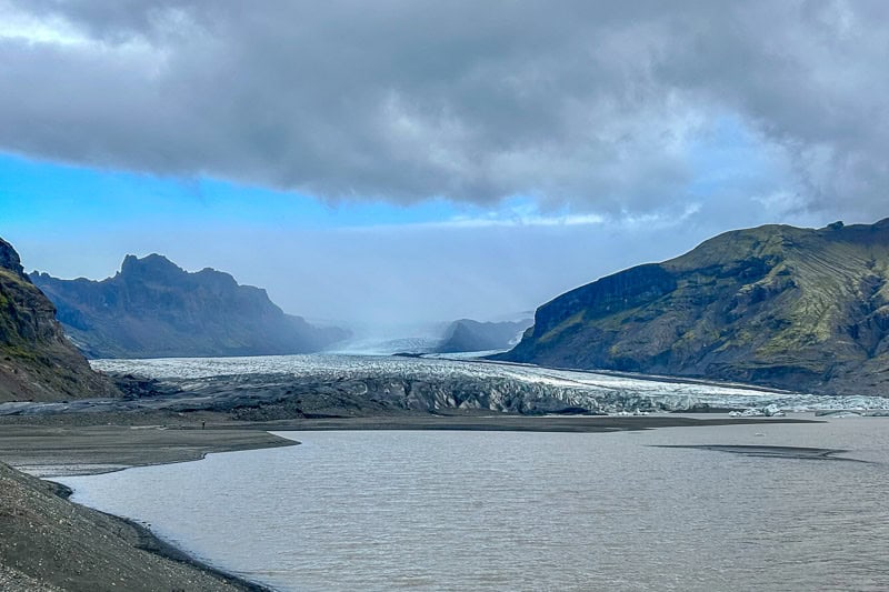 10 Days in Iceland: Our Real Itinerary 22 A glacier flows between rocky, green mountains under a cloudy sky, with a wide river or lake in the foreground—an unforgettable sight during 10 days in Iceland.