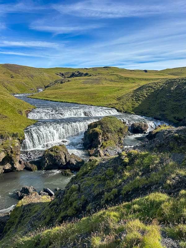 5 Must-See Waterfalls in Iceland 18 A wide, tiered waterfall flows over rocks through a grassy, green landscape under a blue sky with thin clouds. The river bends as it continues through the rolling hills.