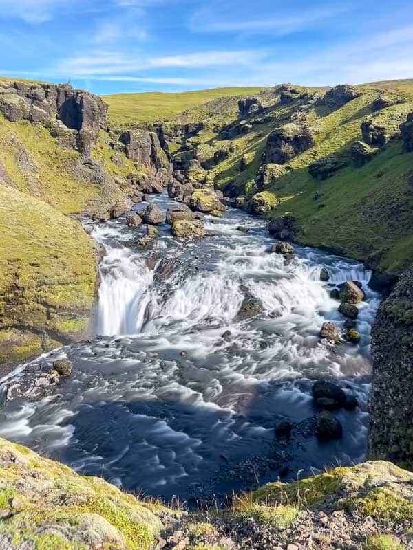 5 Must-See Waterfalls in Iceland 20 A river flows through a rocky, green landscape with small waterfalls and rapids. Steep, grassy hills rise on both sides under a blue sky with scattered clouds.