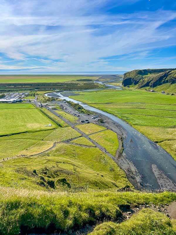 5 Must-See Waterfalls in Iceland 17 A wide, shallow river curves through a green valley with grassy hills, scattered vehicles, and distant cliffs under a blue sky with thin clouds. Fields and a road are visible beside the river.