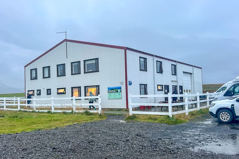 Iceland Campgrounds 14 A two-story white industrial building with several windows, surrounded by a white fence, on a cloudy day. There are people and parked vehicles nearby.