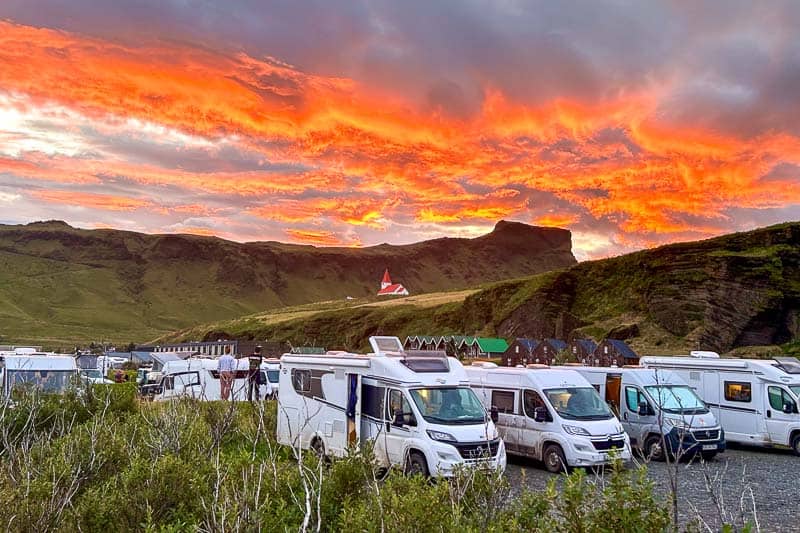 Iceland Campgrounds 7 Several campervans are parked at a campsite with green hills, a church, and dramatic orange clouds at sunset in the background.