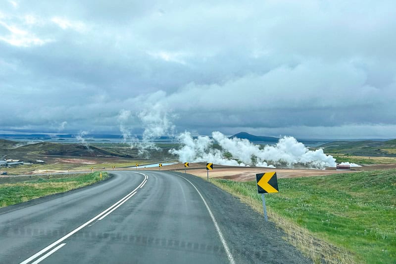 The Diamond Circle: Iceland's Other Scenic Drive 13 A two-lane road on the Diamond Circle curves left through a rural landscape with yellow bend signs. Steam rises from the ground in the distance under a cloudy sky, while green fields and hills frame both sides of the road.