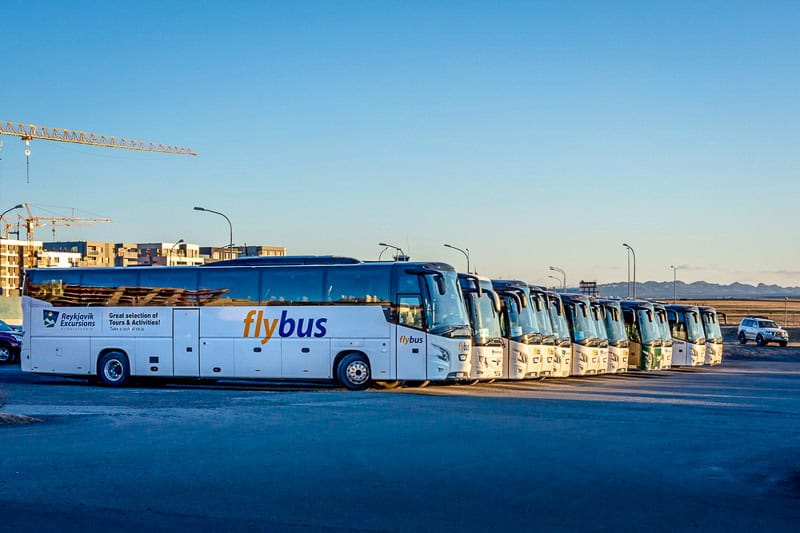 A row of parked Flybus coaches is lined up in a car park under clear blue skies, with some urban buildings and construction cranes visible in the background.