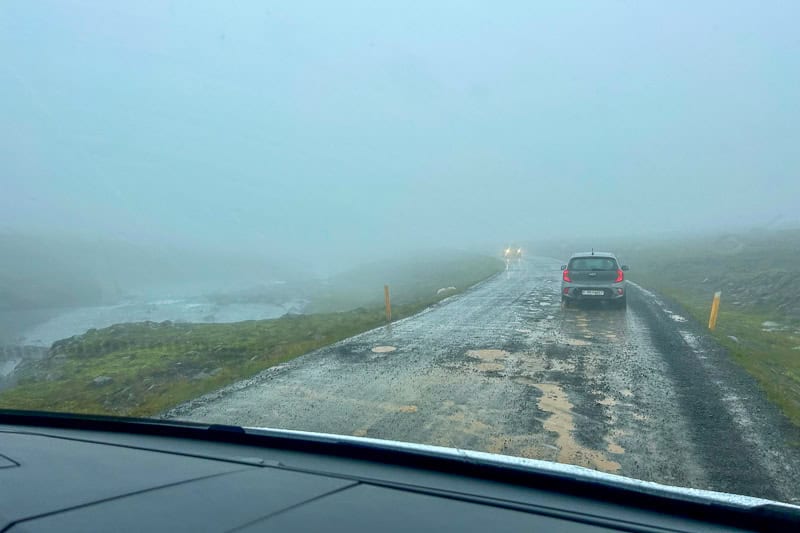 The Diamond Circle: Iceland's Other Scenic Drive 15 A narrow, wet road with potholes and patches winds along the diamond circle, cutting through a foggy landscape. A queue of cars drives ahead with limited visibility due to heavy mist, while mossy ground and rocks border the road.