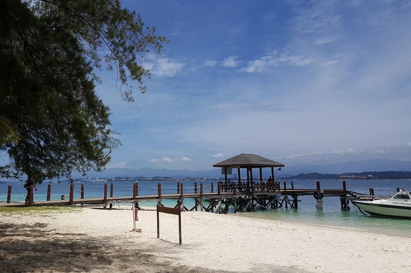 Borneo Group Tour Itinerary (12 Days) - Hosted Small Group Travel 5 A sandy beach with a wooden pier extending into the sea. A small covered pavilion sits on the pier, where people are gathered. A white boat is moored nearby, and trees frame the left side of the image.