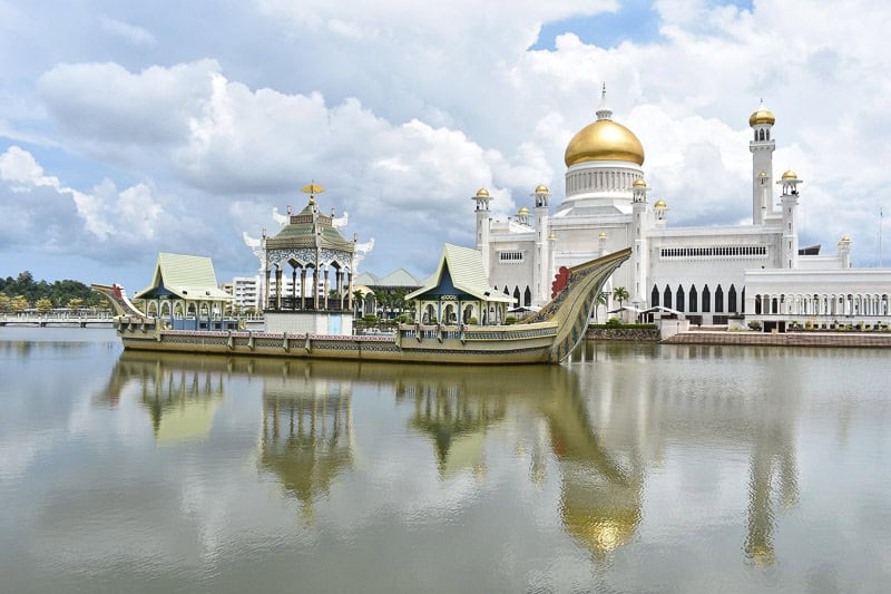 Borneo Group Tour Itinerary (12 Days) - Hosted Small Group Travel 3 A mosque with a golden dome and white walls stands beside a reflective body of water, featuring a ceremonial boat structure in front. The sky is partly cloudy.