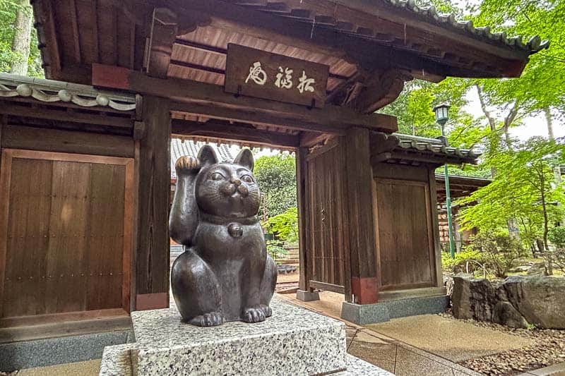 Free Things to Do in Tokyo 1 A large black maneki-neko (beckoning cat) statue sits on a stone plinth in front of a traditional wooden Japanese gate surrounded by trees.