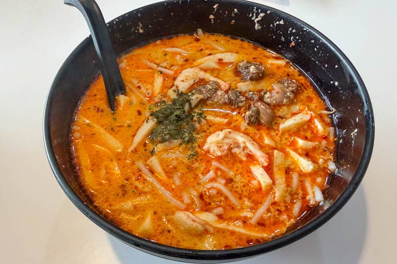 Must-Try Food in Singapore: What to Eat on Your Visit 4 A black bowl filled with spicy orange soup containing noodles, prawns, pieces of meat, and greens, with a black spoon resting inside. The soup appears creamy and is flecked with herbs and seasonings.