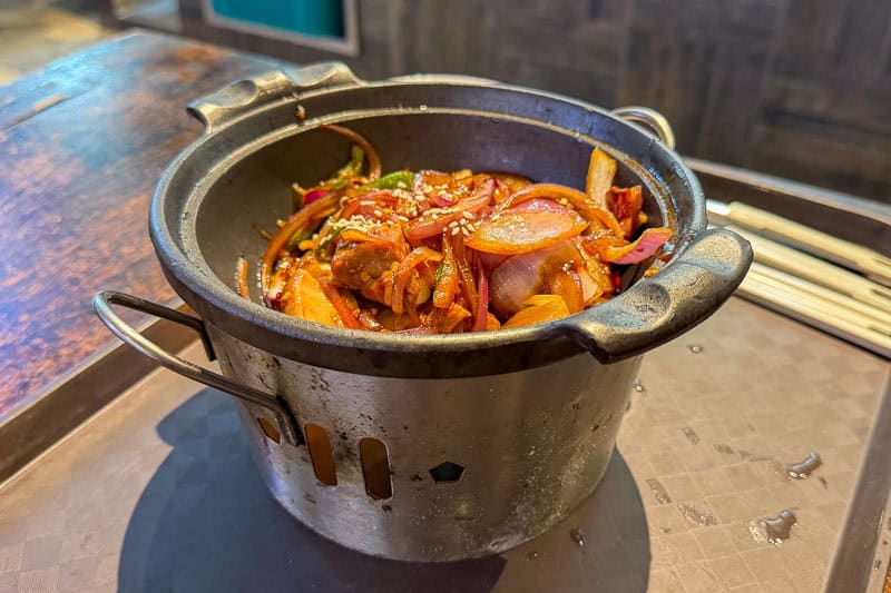 Must-Try Food in Singapore: What to Eat on Your Visit 10 A metal pot filled with stir-fried vegetables, meat, and sauce sits on a brown tray. The dish is garnished with sesame seeds, and chopsticks are placed nearby. The background shows a wooden surface.