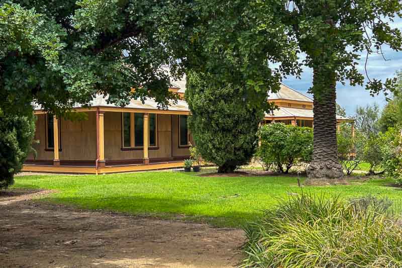 13 Things to Do in Hay, NSW 7 A single-storey house with a wraparound veranda sits behind trees and bushes. The lawn is green and well-kept, with a dirt path in the foreground. Overcast sky is visible in the background.