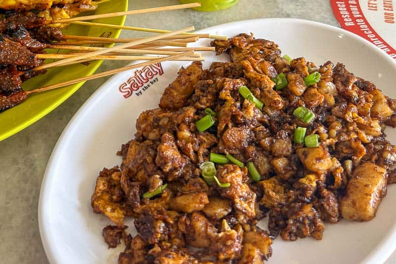 Must-Try Food in Singapore: What to Eat on Your Visit 9 A white plate with stir-fried radish cake pieces garnished with chopped spring onions, next to a yellow plate holding skewered grilled meat. Both dishes are on a light-coloured table.