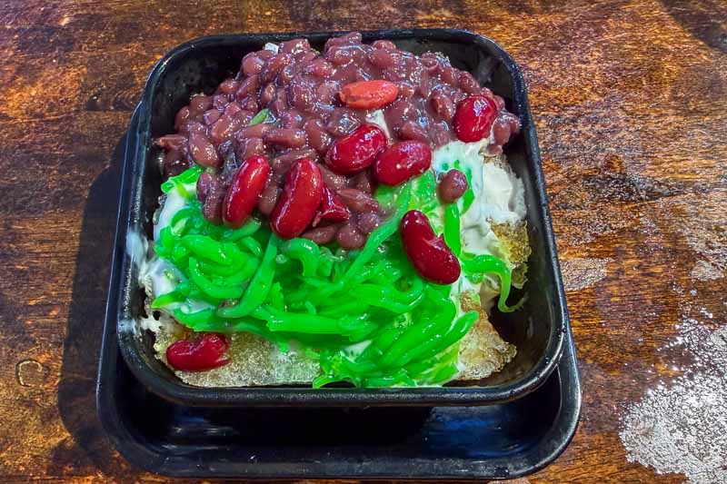 Must-Try Food in Singapore: What to Eat on Your Visit 18 A black square bowl on a wooden surface contains shaved ice topped with red beans, kidney beans, and green pandan jelly strips.