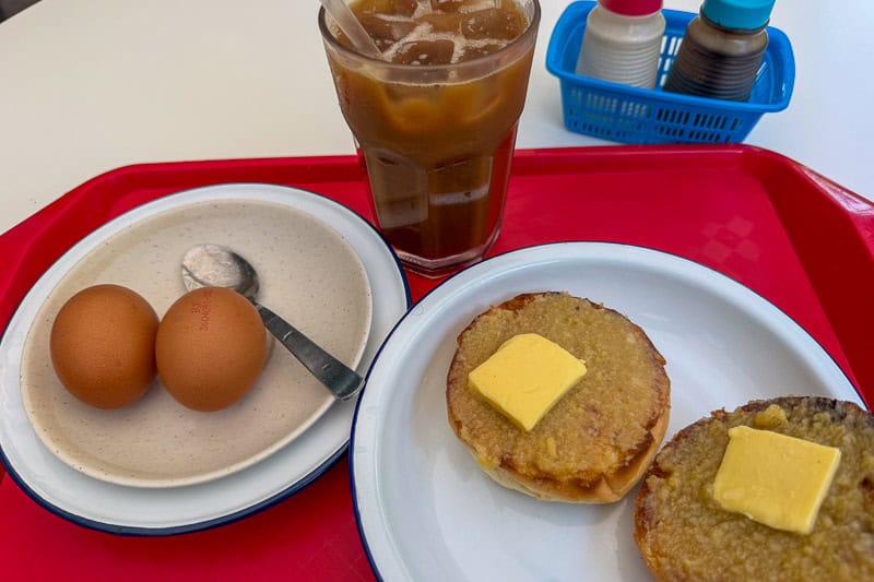 Where to Eat Kaya Toast in Singapore: 9 Places I've Tried 18 A plate with two boiled eggs and a spoon, a glass of iced coffee, and a plate with toasted buns topped with butter, all set on a red tray. Condiment containers are in the background.