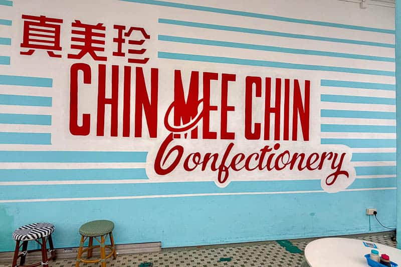 Where to Eat Kaya Toast in Singapore: 9 Places I've Tried 17 Wall mural with large red text reading “CHIN MEE CHIN Confectionery,” flanked by teal horizontal stripes. Chinese characters are above the English text. Two stools and part of a round table are visible below.