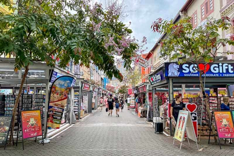 32 Fun Things to do in Singapore on a Budget 5 Pagoda Street, Chinatown, Singapore. People walk down a pedestrian street lined with souvenir shops and trees. Shopfronts display signs and products outside, and colourful decorations hang above the street. The sky is partly cloudy.