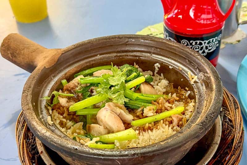 Must-Try Food in Singapore: What to Eat on Your Visit 13 A clay pot filled with rice, pieces of chicken, and green vegetables, garnished with herbs. A red and black container and a yellow drink are in the background on a light blue table.
