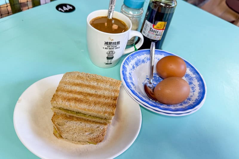 Where to Eat Kaya Toast in Singapore: 9 Places I've Tried 26 A cup of coffee, two soft-boiled eggs in a blue-rimmed bowl, and a plate with two pieces of toasted bread on a light blue table. Bottles of soy sauce and pepper are in the background.