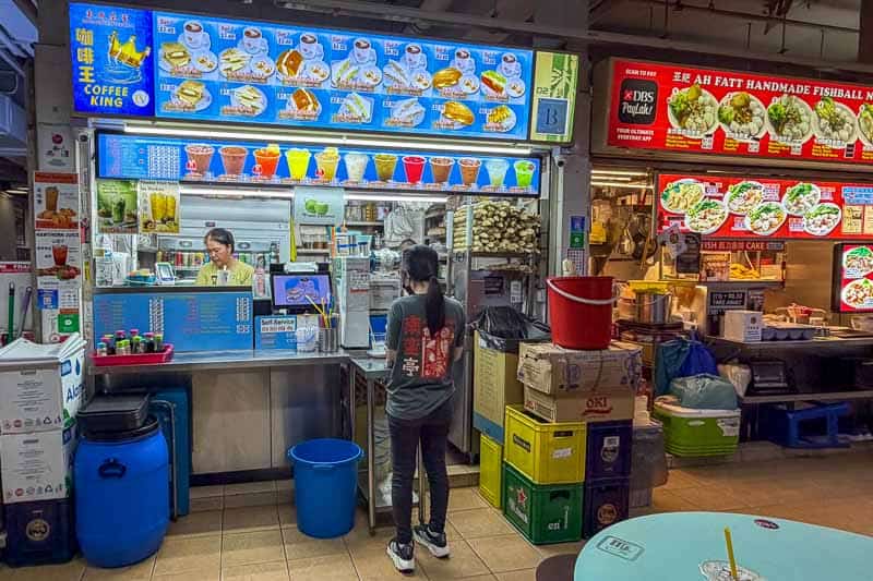 Where to Eat Kaya Toast in Singapore: 9 Places I've Tried 25 A person stands in front of a brightly lit stall with a menu of drinks and food items. The stall is surrounded by containers, crates, and supplies, with another food stall visible to the right.