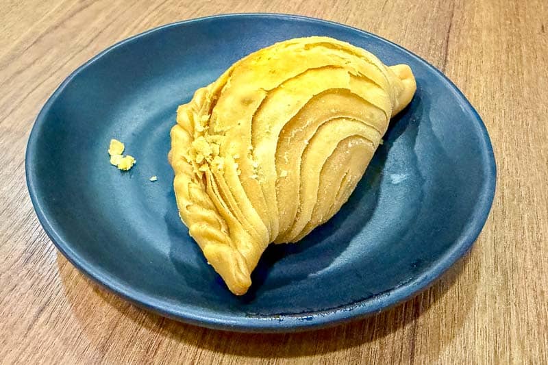 Must-Try Food in Singapore: What to Eat on Your Visit 15 A single golden-brown curry puff with flaky, layered pastry sits on a dark blue plate placed on a wooden table.
