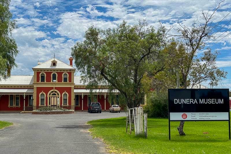 13 Things to Do in Hay, NSW 4 A red two-storey building with arched windows sits behind a sign reading “Dunera Museum.” There are trees and a driveway with parked cars under a partly cloudy blue sky.