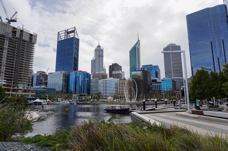 Essential Things to do in Perth on Your First Visit 5 A city skyline with modern high-rise buildings, a harbour with boats, an abstract sculpture near the water, trees, and a cloudy sky above. Construction cranes are visible on the left.