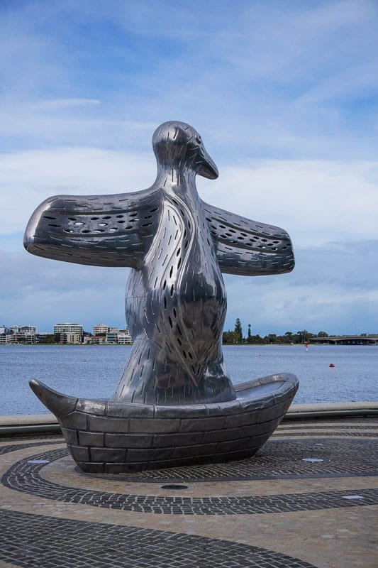 Essential Things to do in Perth on Your First Visit 6 A large metal sculpture of a stylised bird with outstretched wings stands on a circular patterned pavement by a waterfront, with buildings and trees visible across the water under a blue sky.