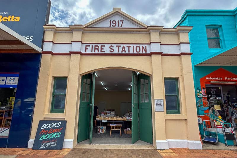 13 Things to Do in Hay, NSW 9 A tan building labelled “1917 Fire Station” with open green doors. Inside, tables display books. A sign outside reads “BOOKS Pre-Loved BOOKS.” Brightly coloured neighbouring shops are visible on either side.
