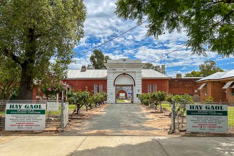 13 Things to Do in Hay, NSW 5 A view of the entrance to Hay Gaol Museum, with a white archway, red brick buildings, and signs on each side displaying visiting hours and information. Trees and bushes line the path leading to the entrance.