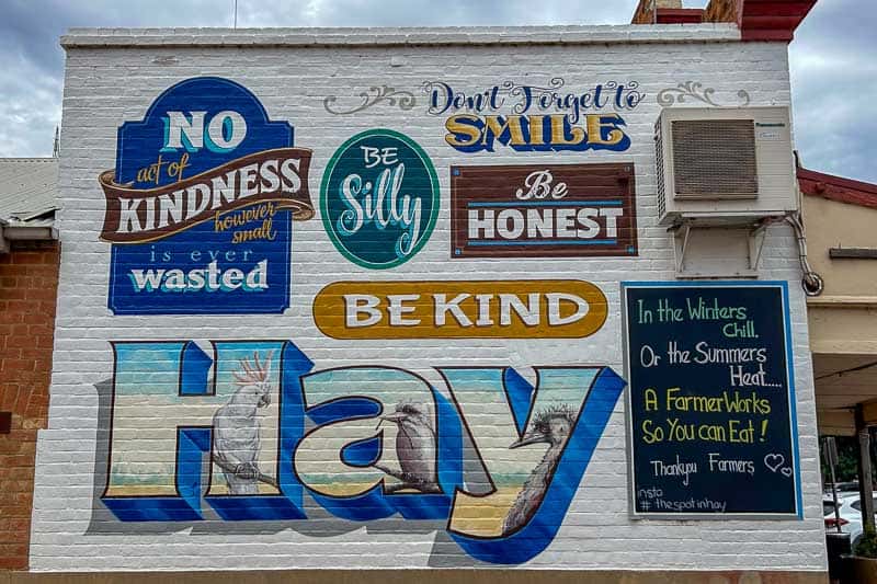 13 Things to Do in Hay, NSW 16 A mural on a brick wall displays messages promoting kindness, honesty, and gratitude for farmers. The artwork features bold and colourful prints, birds, and a blackboard with a note about farmers and food.