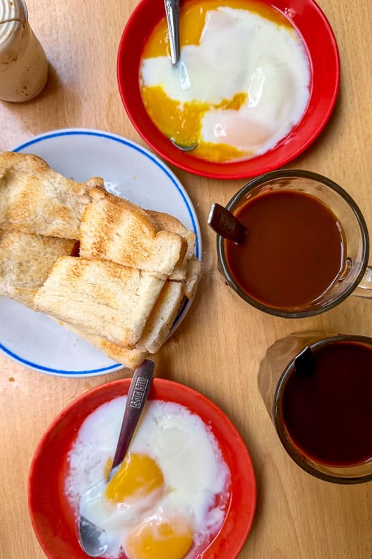 Where to Eat Kaya Toast in Singapore: 9 Places I've Tried 15 A table with two plates of soft-boiled eggs, a plate of toasted bread slices, two mugs of dark coffee, and a glass bottle. Spoons rest in the dishes of eggs and mugs. The setting is indoors on a wooden surface.