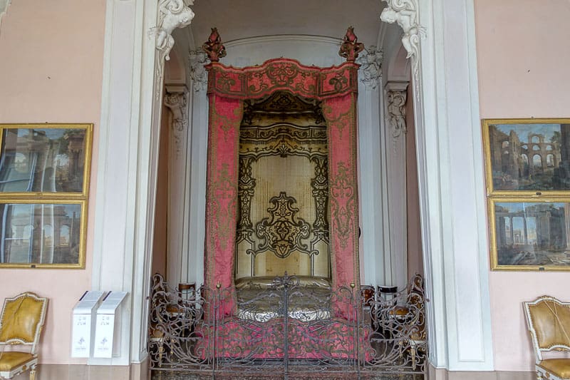 How to Take a Day Trip to the Borromean Islands, Italy 3 Ornate four-poster bed with gold and red canopy, intricate metalwork at the foot, flanked by columns and gilded chairs, set in an elegant room with framed art on pale pink walls.